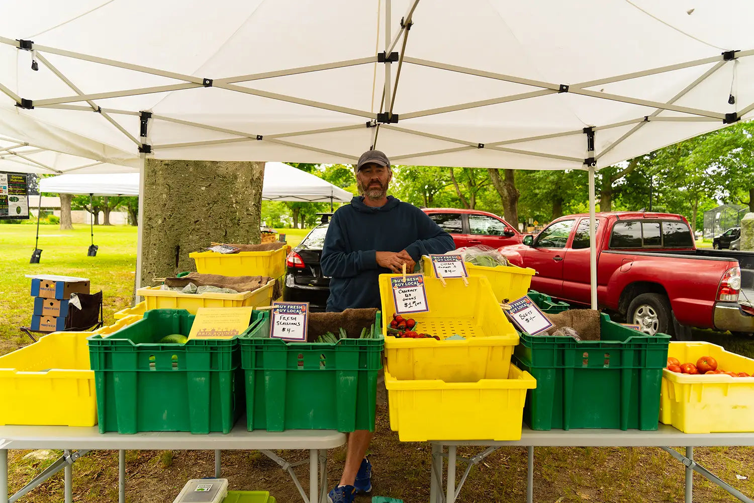 Man standing under a white tent with yellow and green crates at an outdoor market at Coastal Foodshed