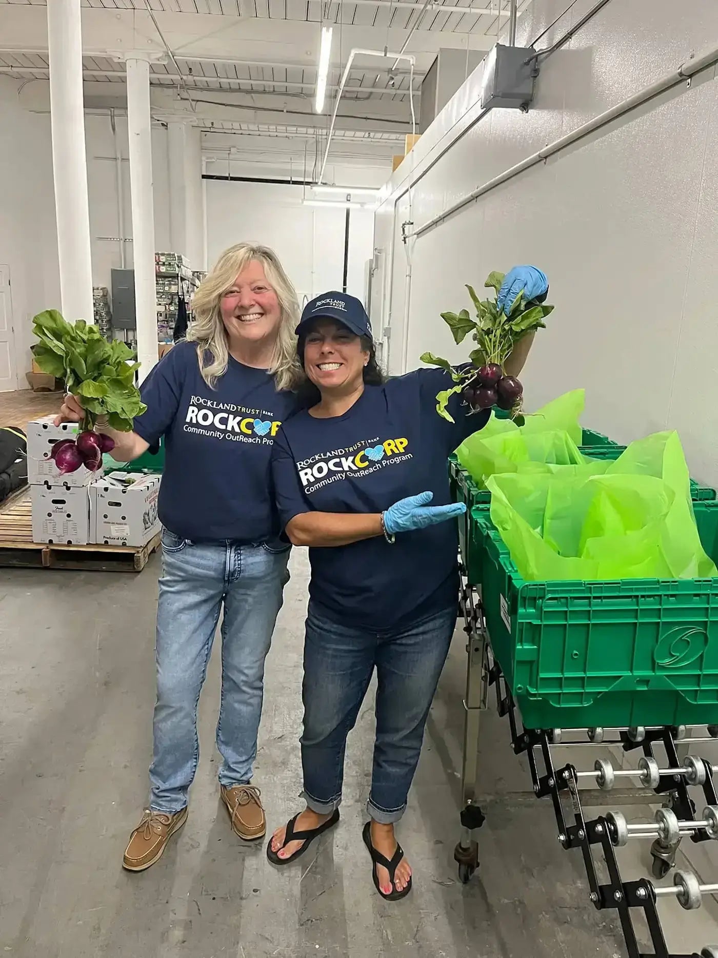 two HIP Delivery Program workers holding fresh produce next to green crates & bags