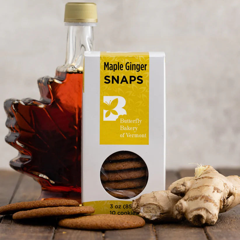 Box of Maple Ginger Snaps with a bottle of maple syrup and ginger root on a wooden surface.