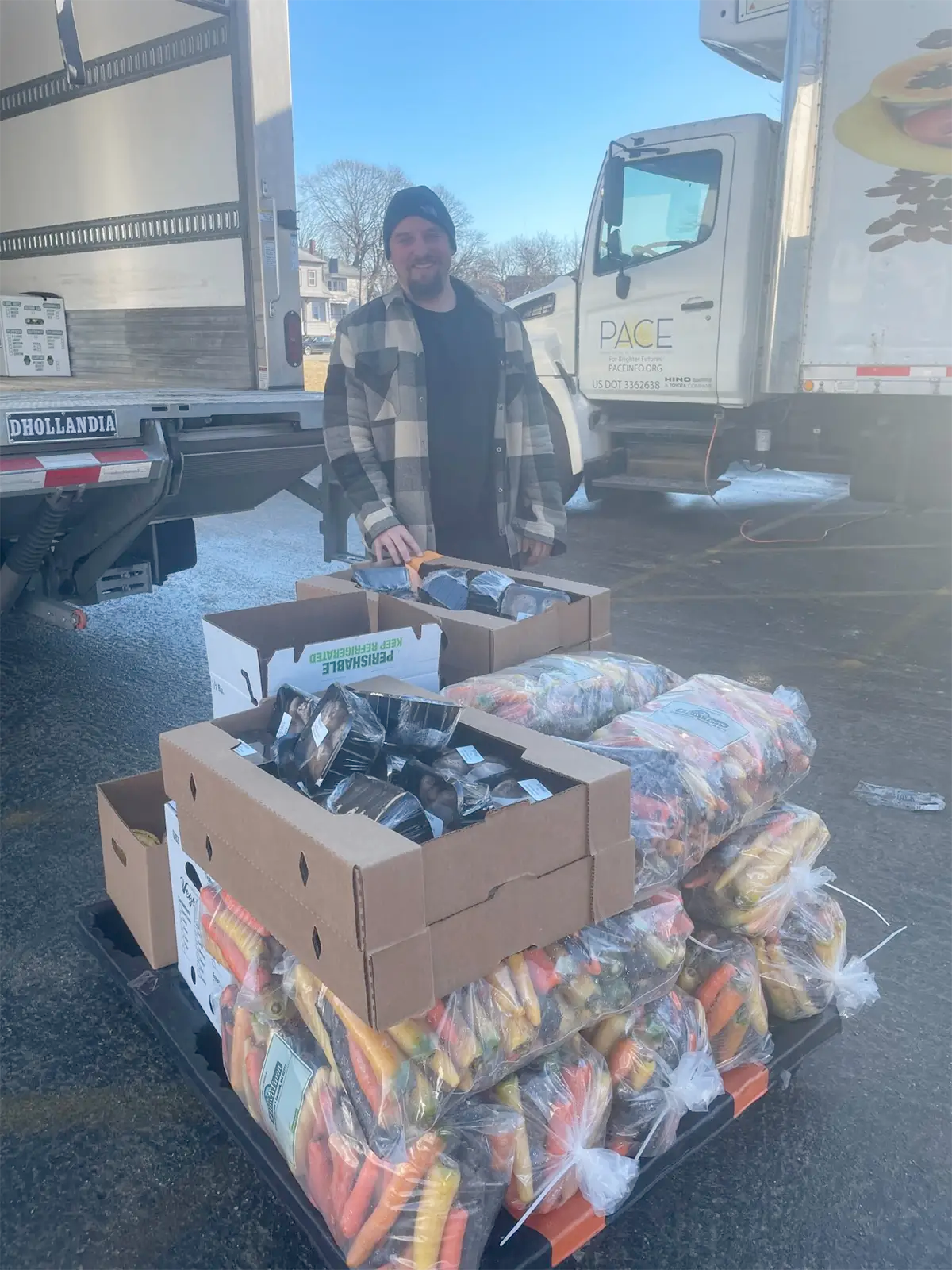 Coastal Foodshed worker standing behind pallet of assorted produce, trucks in background